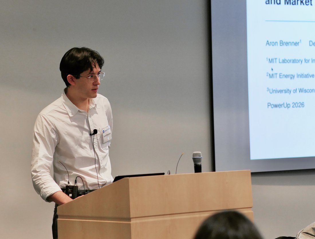 Aron Brenner speaking at a podium beside a projected title slide reading Strategic Spatial Load Shifting and Market Efficiency, presented at PowerUp 2026.